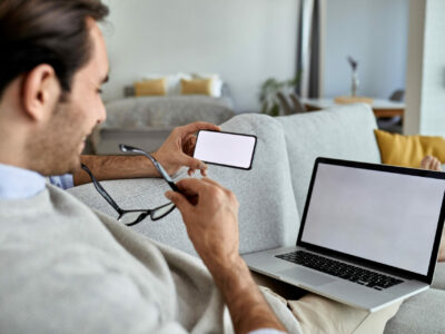 Close-up of a man reading text message on mobile phone while working laptop at home. Copy space.